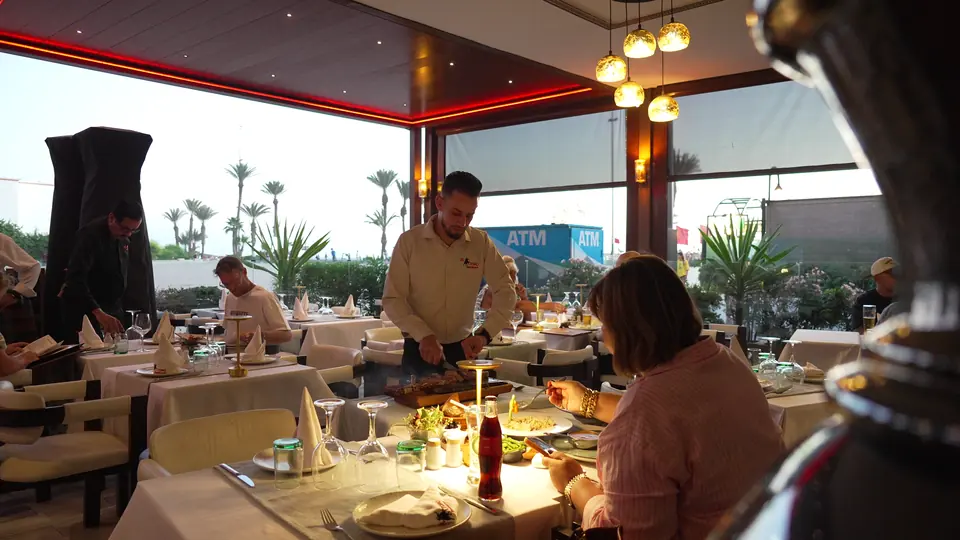 Un serveur sert une cliente à une table près des baies, au coucher du soleil